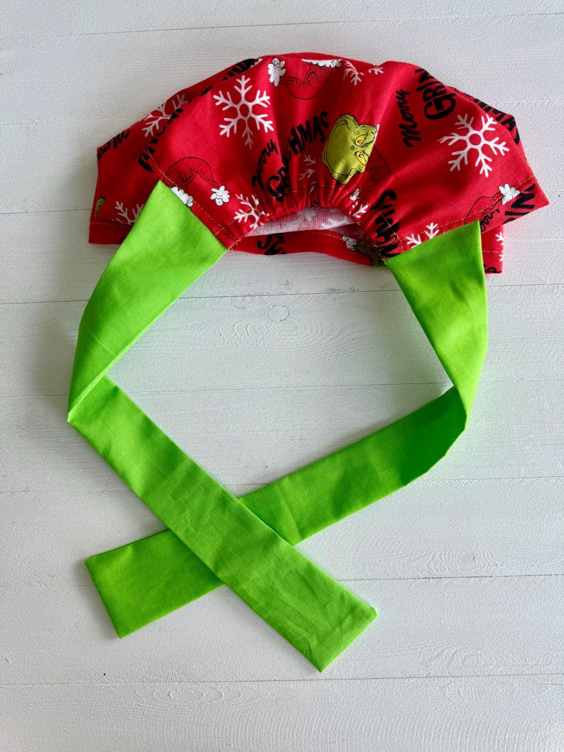 May include: A red fabric scrub cap with a green tie. The cap features white snowflakes and the text "Merry Grinchmas" with a cartoon Grinch face. The tie is a bright green color.