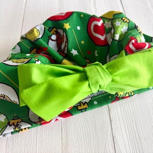 May include: A green fabric headband with a bright green bow. The headband is patterned with Christmas ornaments, stars, and cartoon characters on a green background. The bow is a solid, bright green color.