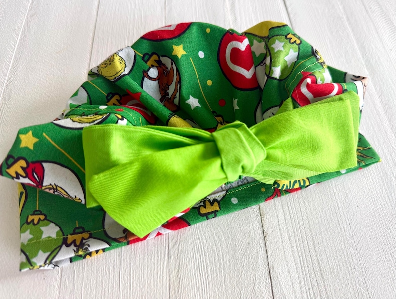 May include: A green fabric headband with a bright green bow. The headband is patterned with Christmas ornaments, stars, and cartoon characters on a green background. The bow is a solid, bright green color.