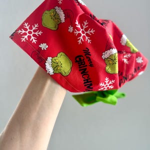 May include: Red fabric headband with a Grinch and snowflake pattern. The text "Merry Grinchmas" is printed on the fabric. A green bow is attached to the headband.