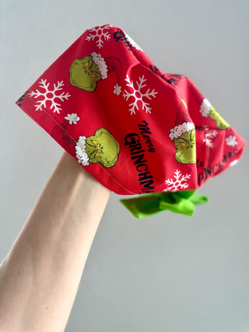 May include: Red fabric headband with a Grinch and snowflake pattern. The text "Merry Grinchmas" is printed on the fabric. A green bow is attached to the headband.