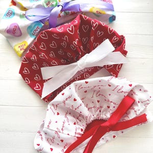May include: Three Valentine's Day themed scrub caps. One is white with candy heart graphics and a purple ribbon. Another is red with white hearts and a white ribbon. The third is white with red "I love you" text and a red ribbon.