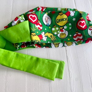 May include: A green fabric headband with a Grinch-themed pattern featuring the Grinch, ornaments, and the word "GRINCH". The headband has a bright green tie.