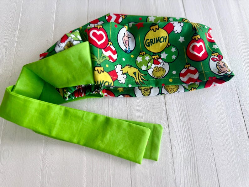 May include: A green fabric headband with a Grinch-themed pattern featuring the Grinch, ornaments, and the word "GRINCH". The headband has a bright green tie.