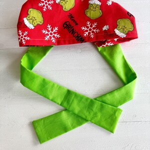 May include: A red surgical scrub cap with a green tie. The cap features a pattern of the Grinch, snowflakes, and the text "Merry Grinchmas". The tie is a solid bright green.