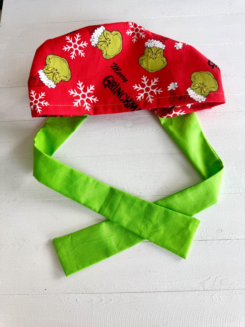 May include: A red surgical scrub cap with a green tie. The cap features a pattern of the Grinch, snowflakes, and the text "Merry Grinchmas". The tie is a solid bright green.