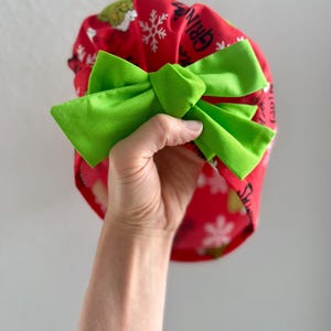 May include: A red scrub cap with a green bow. The cap features a pattern of white snowflakes and a cartoon character. The word "GRINCH" is printed on the cap.