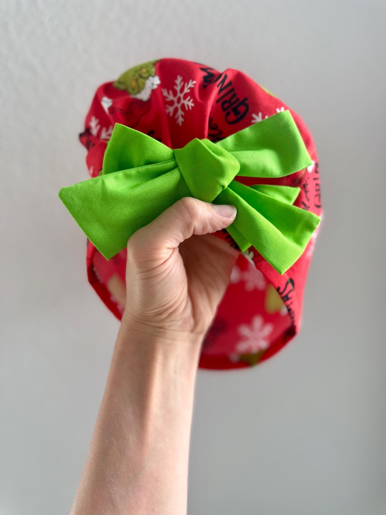 May include: A red scrub cap with a green bow. The cap features a pattern of white snowflakes and a cartoon character. The word "GRINCH" is printed on the cap.