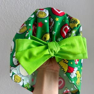 May include: A green fabric scrub cap with a bright green bow. The cap features a pattern of cartoon characters, red stars, and ornaments on a green background. The bow is a solid, vibrant green.