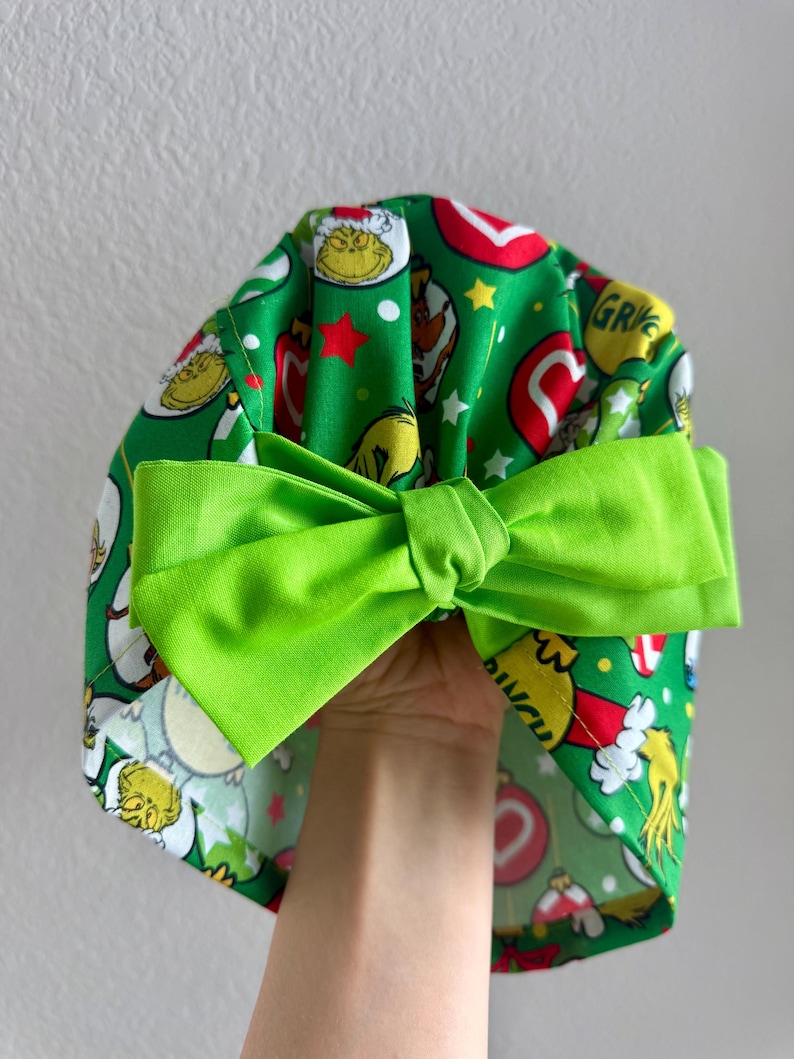 May include: A green fabric scrub cap with a bright green bow. The cap features a pattern of cartoon characters, red stars, and ornaments on a green background. The bow is a solid, vibrant green.