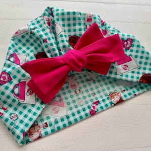 May include: A turquoise and white gingham fabric turban with a pink bow. The fabric has a repeating pattern of small pink purses and hearts.