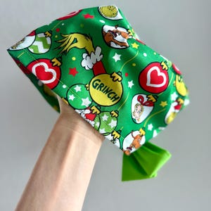 May include: Green fabric head covering with a Grinch-themed pattern. The design includes Grinch characters, Christmas ornaments, and the word "Grinch" on a yellow ornament. A bright green bow is attached.