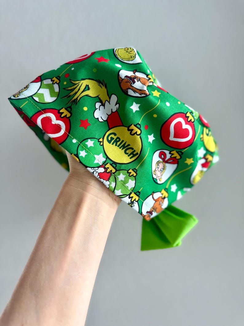 May include: Green fabric head covering with a Grinch-themed pattern. The design includes Grinch characters, Christmas ornaments, and the word "Grinch" on a yellow ornament. A bright green bow is attached.