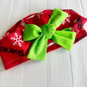 May include: A red fabric headband with a large, bright green bow. The headband features white snowflakes and the word "GRINCHMAS" in black lettering. The bow is centered on the headband, creating a festive holiday accessory.