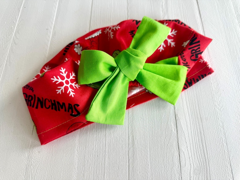 May include: A red fabric headband with a large, bright green bow. The headband features white snowflakes and the word "GRINCHMAS" in black lettering. The bow is centered on the headband, creating a festive holiday accessory.