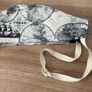 May include: A white fabric scrub hat with a black and white vintage world map print. The hat has a long white tie that can be tied around the neck. The text on the map reads "Map of the World".