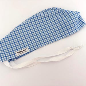 May include: A blue and white gingham patterned scrub cap featuring a white fabric label with the black text SCRUBS AHOY Handmade in Texas. The cap includes two long white fabric ties extending from the base for securing the head covering.