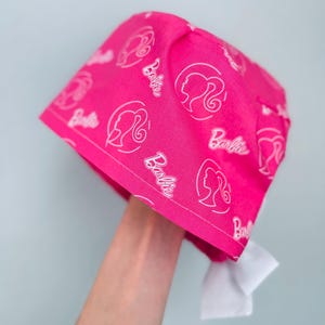 May include: Pink surgical scrub hat with a white Barbie logo pattern. The hat has a white tie at the back.