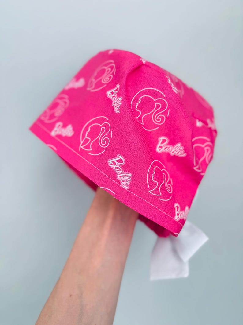 May include: Pink surgical scrub hat with a white Barbie logo pattern. The hat has a white tie at the back.