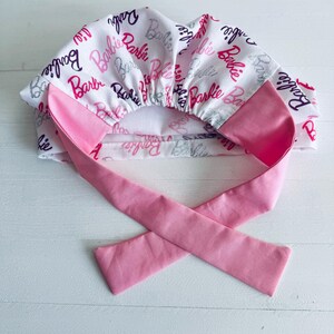 May include: A pink and white surgical scrub hat with a tie. The hat is covered in a repeating pattern of the word "Barbie" in pink, purple, and silver glitter.