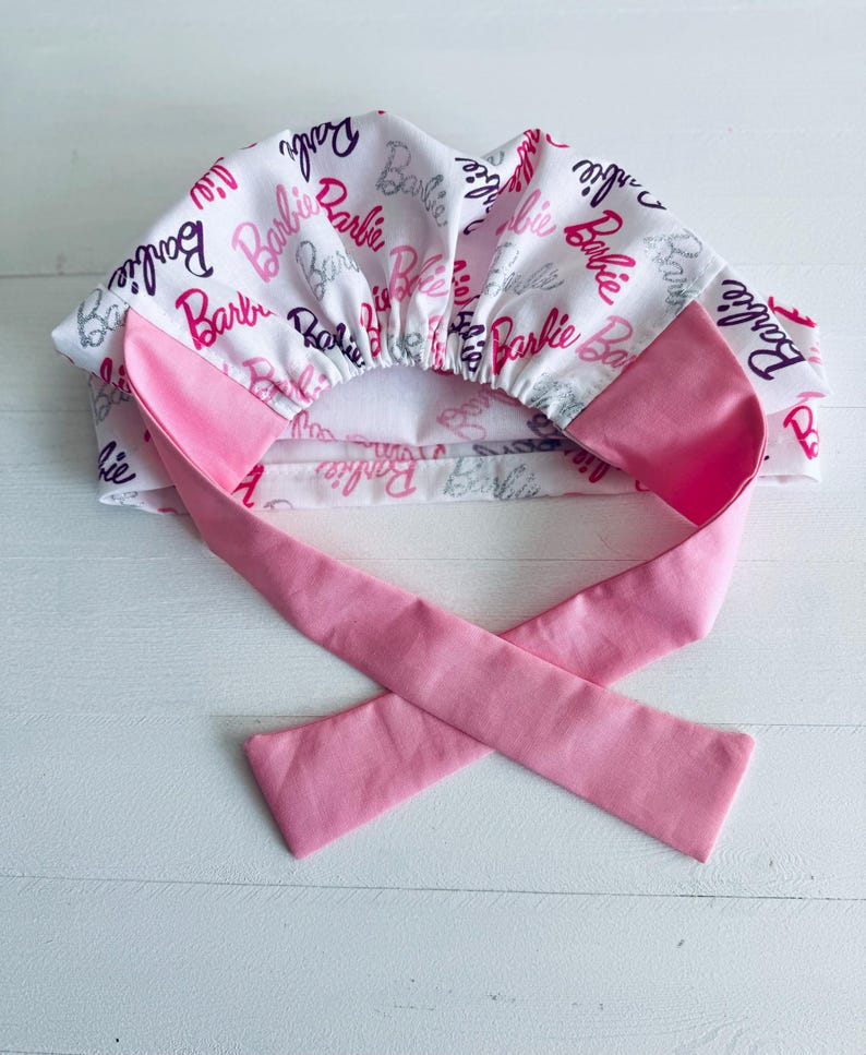 May include: A pink and white surgical scrub hat with a tie. The hat is covered in a repeating pattern of the word "Barbie" in pink, purple, and silver glitter.
