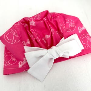 May include: Pink fabric shower cap with a white bow and a repeated pattern of the Barbie logo and silhouette.