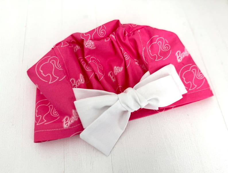 May include: Pink fabric shower cap with a white bow and a repeated pattern of the Barbie logo and silhouette.