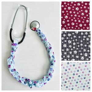 May include: A stethoscope with a blue and purple paw print patterned fabric cover. The stethoscope is on a white surface. The fabric cover is a stretchy material that can be used to hold the stethoscope in place.