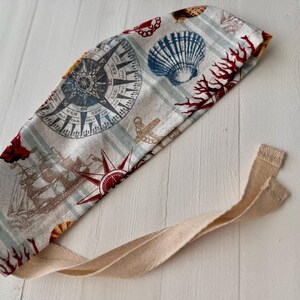 May include: A beige fabric headband with a nautical theme. The headband features a compass, seashells, coral, and a ship. The headband has a long beige tie.