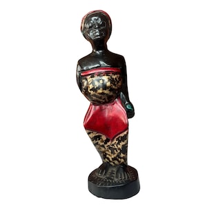 Puede incluir: Una figurita de madera de una persona con un cuerpo negro, una falda de estampado rojo y negro y una piedra verde en la mano.