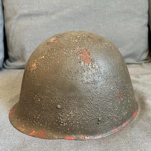Puede incluir: Un casco militar verde oliva con una superficie texturizada y ribete rojo. El casco muestra signos de desgaste, con zonas de pérdida de pintura y óxido. Es un artículo vintage.