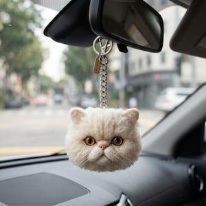May include: A cream-colored, fluffy cat head keychain hanging from a silver-toned metal chain and lobster clasp. The cat features textured, wool-like fur, prominent amber-colored eyes, a small pink nose, and long white whiskers, suspended from a rearview mirror.