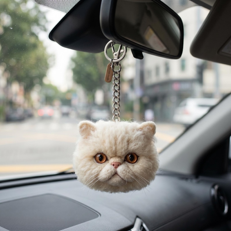 May include: A cream-colored, fluffy cat head keychain hanging from a silver-toned metal chain and lobster clasp. The cat features textured, wool-like fur, prominent amber-colored eyes, a small pink nose, and long white whiskers, suspended from a rearview mirror.