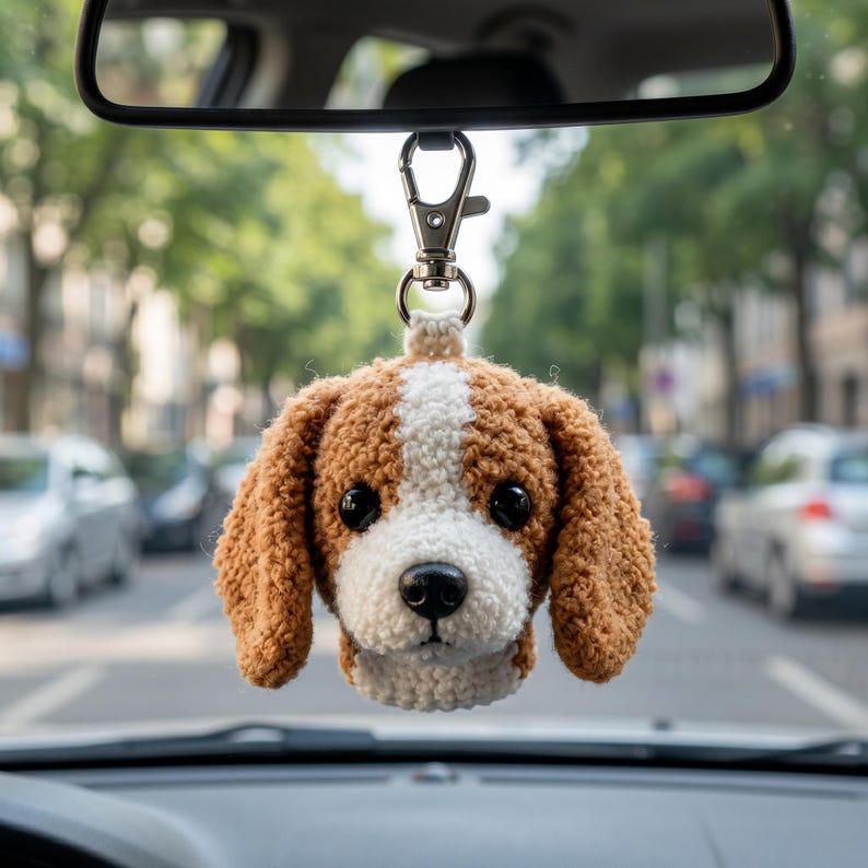 May include: A crocheted beagle dog head ornament hanging from a silver-toned metal lobster clasp. The plush toy features brown and white yarn, black plastic eyes, and a black nose, suspended from a car rearview mirror for interior decoration.