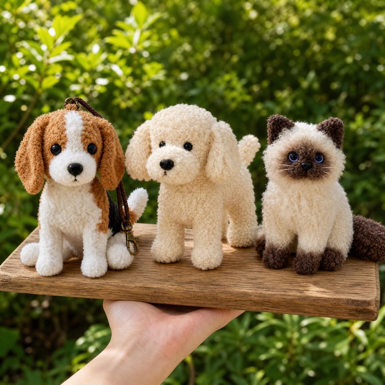 May include: Three small, crocheted animal figurines: a brown and white beagle with a dark brown lanyard, a cream-colored poodle, and a Siamese cat with dark brown ears and tail. The plush toys are arranged in a row on a rectangular wooden board.
