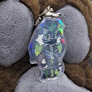 May include: A holographic keychain featuring a cartoon character with green hair, a black and white striped tail, and a green and black outfit. The character has a green and black eye and a black nose.