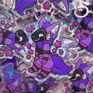 May include: A collection of holographic keychains featuring a purple and black cartoon character with a stylized animal head and striped body. The keychains are surrounded by pink star-shaped charms.