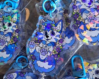 May include: Multiple blue and white cartoon fox keychains with flower crowns and blue clasps. The keychains are in clear plastic packaging. The fox has purple eyes and is surrounded by purple and white flowers.