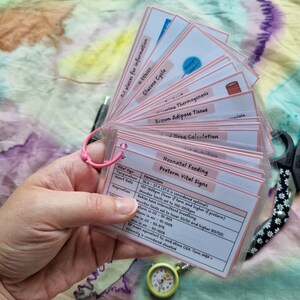 May include: A set of flashcards held together by a pink ring, with topics like "Glucose Cycle" and "Neonatal Feeding." The cards have pink borders and are held near a pair of daisy-patterned scissors and a small medical watch.