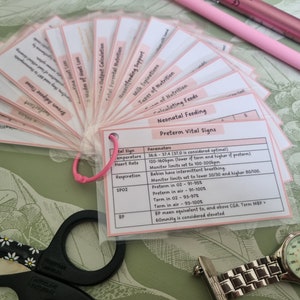 May include: A fan of flashcards with pink tabs, detailing neonatal feeding and preterm vital signs. The cards are held together with a pink cord. A pair of scissors, a watch, and a pink pen are also visible.
