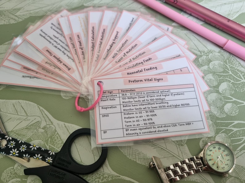May include: A fan of flashcards with pink tabs, detailing neonatal feeding and preterm vital signs. The cards are held together with a pink cord. A pair of scissors, a watch, and a pink pen are also visible.