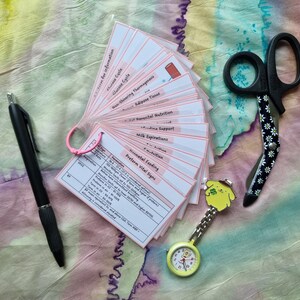 May include: A collection of flashcards with medical information, a black pen, a pair of daisy-patterned scissors, and a yellow nurse's watch with a cartoon character. The flashcards are held together with a pink ring.