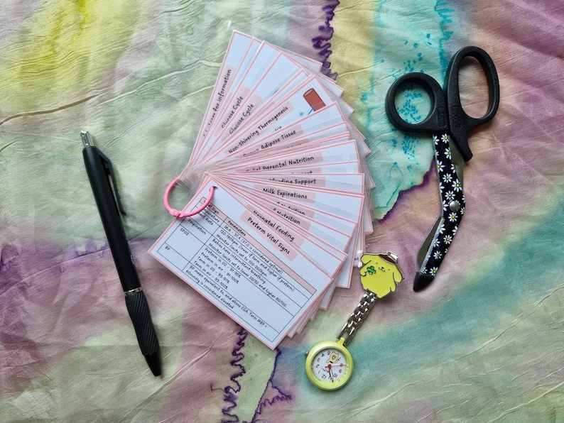 May include: A collection of flashcards with medical information, a black pen, a pair of daisy-patterned scissors, and a yellow nurse's watch with a cartoon character. The flashcards are held together with a pink ring.