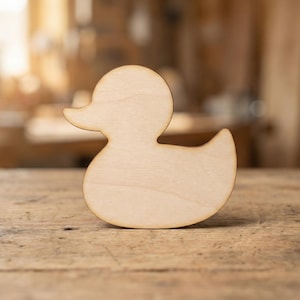 May include: A wooden duck-shaped cutout with a smooth, pale surface. The duck has a rounded body, a small beak, and a curved tail. The cutout is positioned on a wooden surface, with a blurred background.