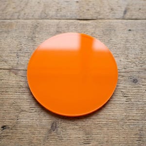 Può includere: Un sottobicchiere circolare arancione brillante con finitura lucida. Il sottobicchiere è posto su uno sfondo di legno rustico. Questo design semplice è adatto a vari stili di arredamento.