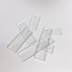 Puede incluir: Un conjunto de etiquetas rectangulares transparentes de acrílico con esquinas redondeadas y un pequeño agujero en la parte superior para colgar. Las etiquetas están dispersas sobre una superficie blanca. Ideal para manualidades o etiquetado.