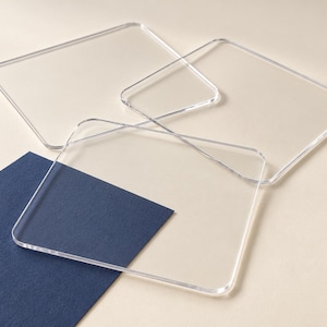 Könnte beinhalten: Drei quadratische Untersetzer aus klarem Acryl mit abgerundeten Ecken, die überlappend angeordnet sind. Die transparenten Untersetzer haben glatte, polierte Kanten und eine flache Oberfläche und liegen teilweise auf einer dunkelblauen rechteckigen Karte.