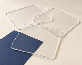 May include: Three clear, square acrylic coasters with rounded corners arranged in an overlapping pattern. The transparent coasters have smooth, polished edges and a flat surface, partially resting on a dark blue rectangular card.
