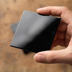 Puede incluir: Un objeto cuadrado, negro y brillante se sostiene en una mano. El objeto refleja la luz, creando reflejos en su superficie. Los bordes parecen afilados y bien definidos. Otro objeto similar es ligeramente visible en el fondo.