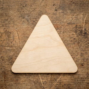 Op de afbeelding: Een lichtgekleurde, houten driehoek met afgeronde hoeken. De driehoek staat tegen een getextureerde, bruine houten achtergrond. De vorm is eenvoudig en geometrisch, geschikt voor knutselen of decoratieve doeleinden.
